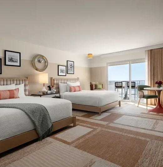 A modern hotel room with two queen beds, wall art, a round table with chairs, a bench, and a large window with curtains leading to a balcony overlooking the ocean.
