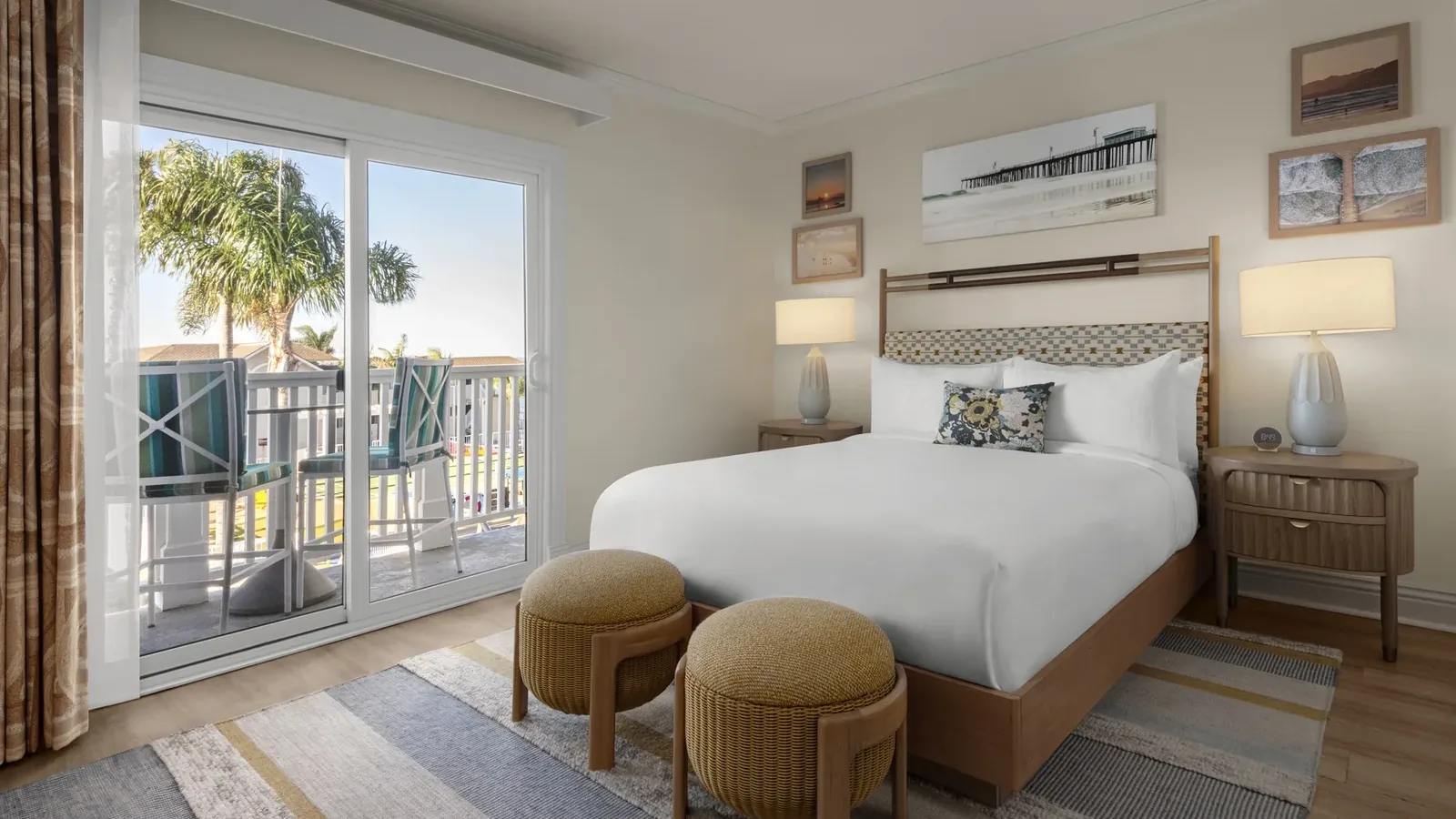 A bright hotel room with a neatly made bed, two round ottomans at the foot, nightstands with lamps, coastal artwork on the walls, and a sliding glass door leading to a balcony with palm trees outside.