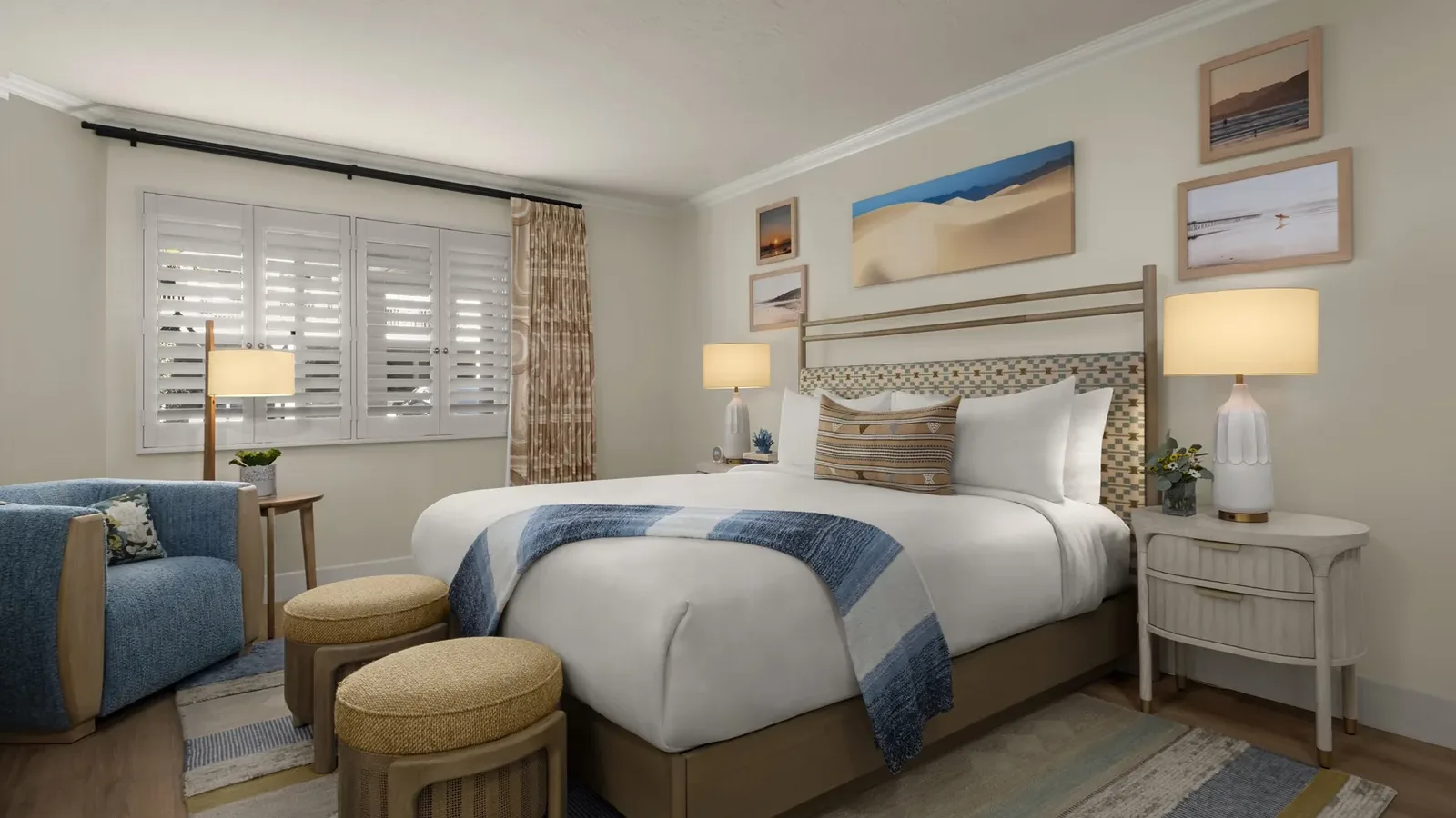 A cozy, modern bedroom with a large bed, blue and beige accents, two round ottomans, a blue armchair, two nightstands with lamps, wall art above the bed, and sunlight streaming through white shutters.