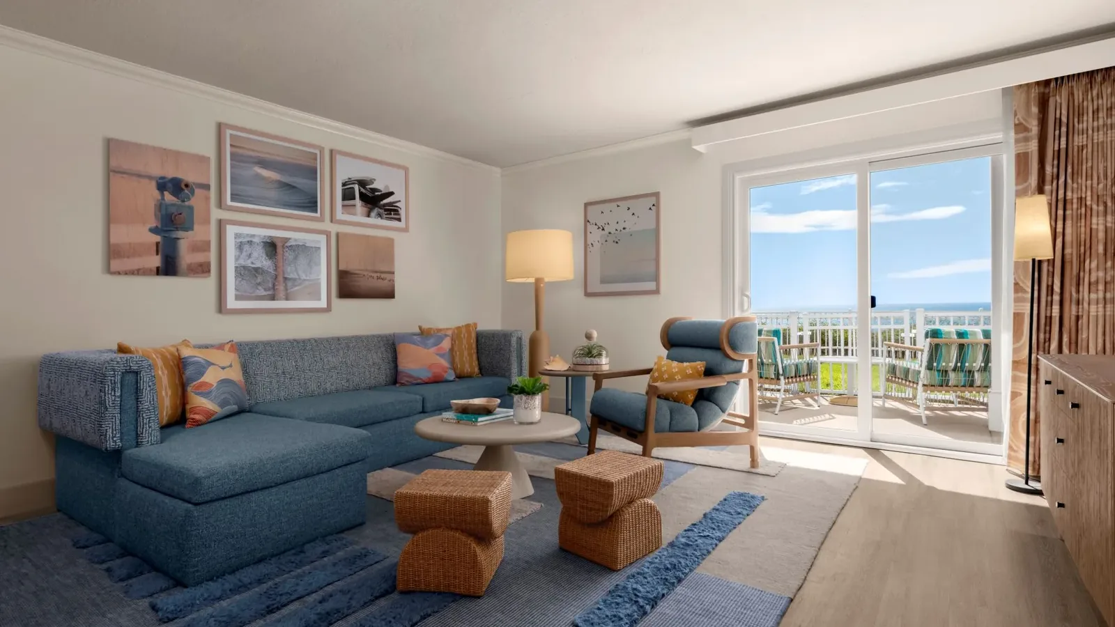 A bright living room with a blue sectional sofa, artwork on white walls, a round coffee table, armchair, woven stools, and a rug; sliding glass doors lead to a balcony with ocean views.