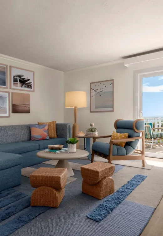 A bright living room with a blue sectional sofa, artwork on white walls, a round coffee table, armchair, woven stools, and a rug; sliding glass doors lead to a balcony with ocean views.