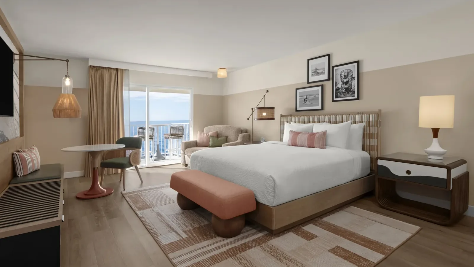 A modern hotel room with a large bed, a nightstand, a lamp, a small dining table with two chairs, an armchair, and a balcony overlooking the ocean. The decor features warm, neutral tones and framed artwork.