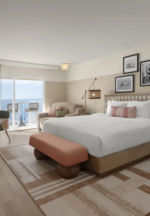A modern hotel room with a large bed, a nightstand, a lamp, a small dining table with two chairs, an armchair, and a balcony overlooking the ocean. The decor features warm, neutral tones and framed artwork.