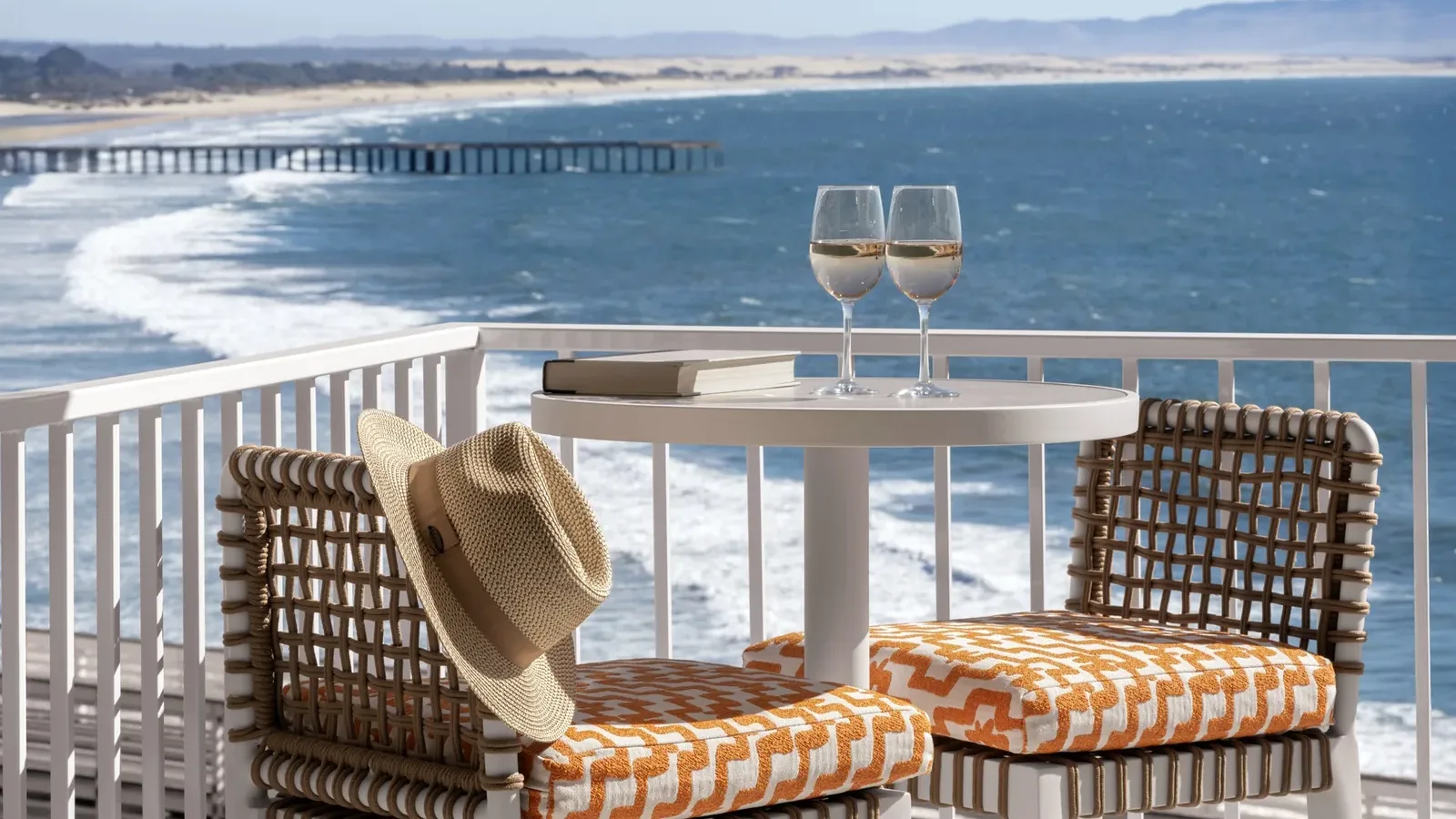 Two chairs with orange patterned cushions and a table with two glasses of white wine, books, and a straw hat overlook an ocean view with waves, a sandy beach, and a distant pier.