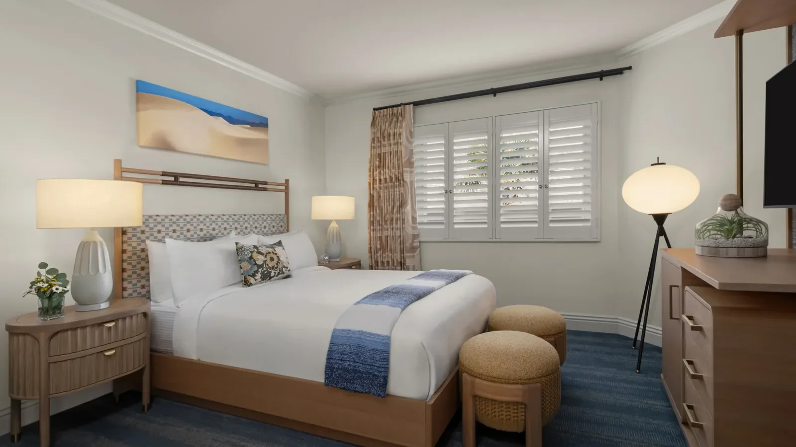 A cozy hotel room with a neatly made queen bed, wooden furniture, two round ottomans at the foot of the bed, blue accents, two bedside lamps, a globe floor lamp, a TV, and a window with white shutters.