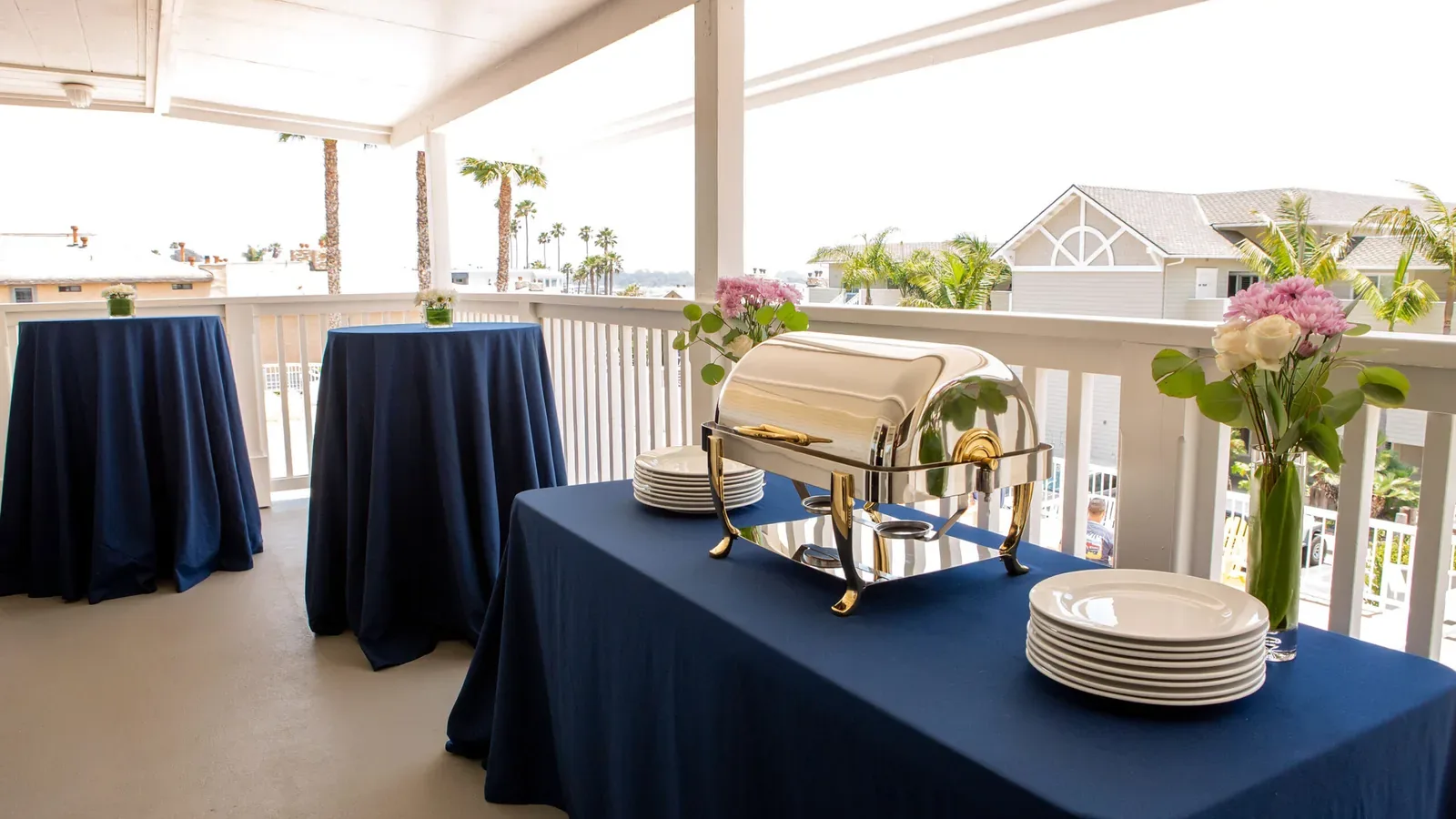 A covered outdoor patio setup with blue tablecloth-covered tables, a chafing dish, stacked white plates, and vases of flowers; palm trees and houses are visible in the background.