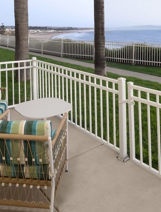 Two cushioned chairs and a small table sit on a patio with white fencing, overlooking a grassy area, palm trees, a coastal path, and the ocean in the background.