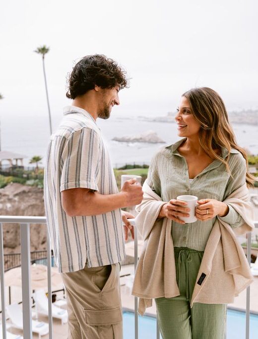 A man and woman stand on a balcony overlooking the ocean, both holding coffee mugs and smiling at each other. The background features palm trees, cliffs, and a cloudy sky.