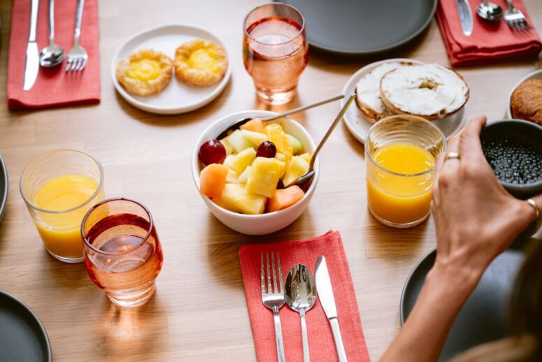 A breakfast table set with plates, glasses of orange juice, a bowl of mixed fruit, pastries, and bagels with cream cheese. Utensils rest on red napkins, and a person’s hand holds a black cup.