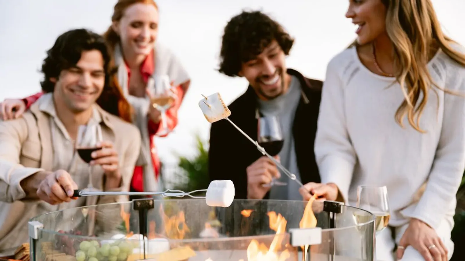 Four friends sit around an outdoor fire pit, smiling and toasting marshmallows on skewers while holding glasses of red wine. There is a platter of fruit and cheese on the table next to the fire.