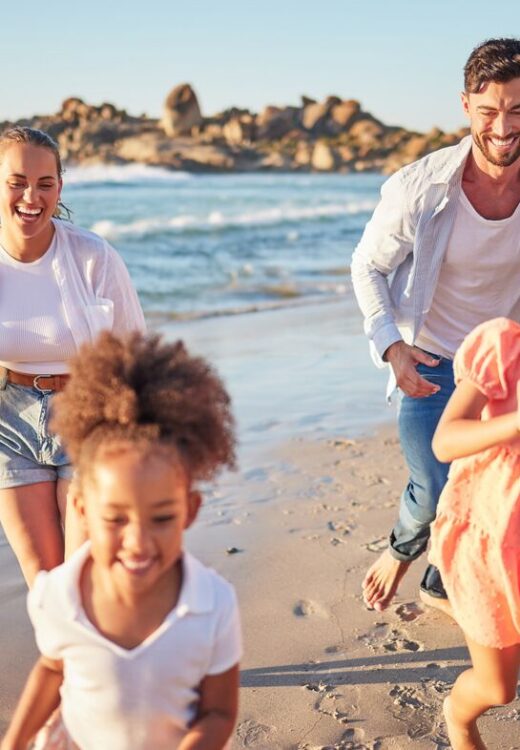 A smiling family of four, two adults and two young children, run and play together along a sunny beach with waves and rocky cliffs in the background. Everyone looks happy and carefree.