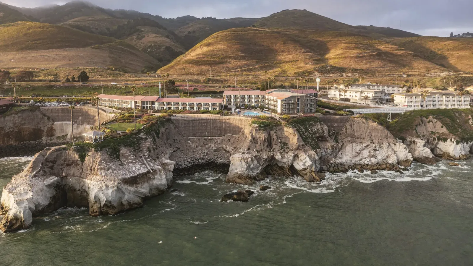 A coastal hotel complex sits atop rugged cliffs overlooking the ocean, with waves crashing below and rolling green hills in the background under a cloudy sky.