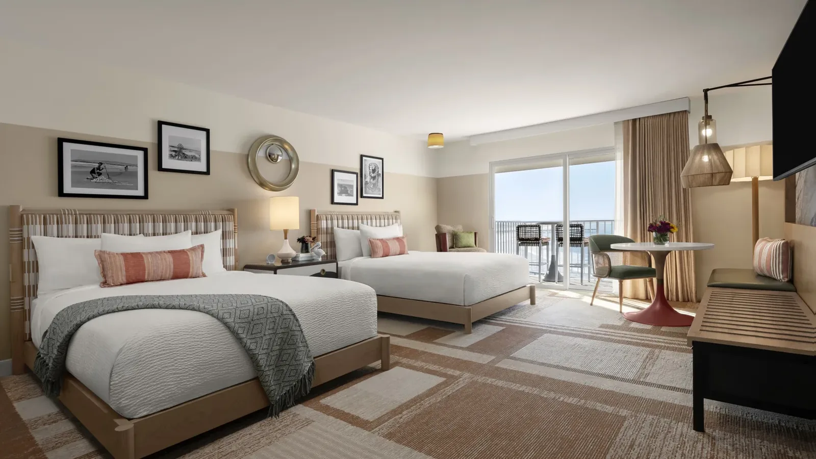 Modern hotel room with two double beds, decorative pillows, framed artwork on the walls, a round table with two chairs, a bench, and a large window with curtains leading to a balcony with an ocean view.
