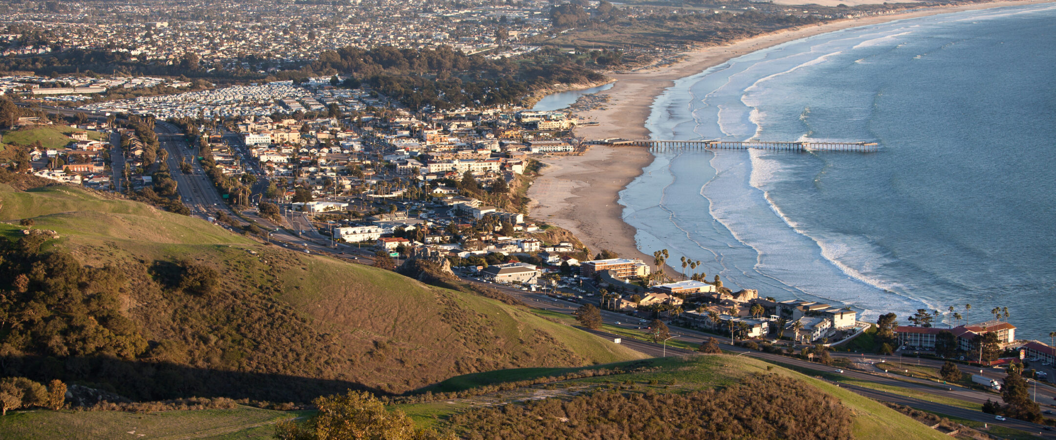 A coastal city with a long pier extends into the ocean, bordered by a wide sandy beach and a stretch of green hills in the foreground, under a clear sky.
