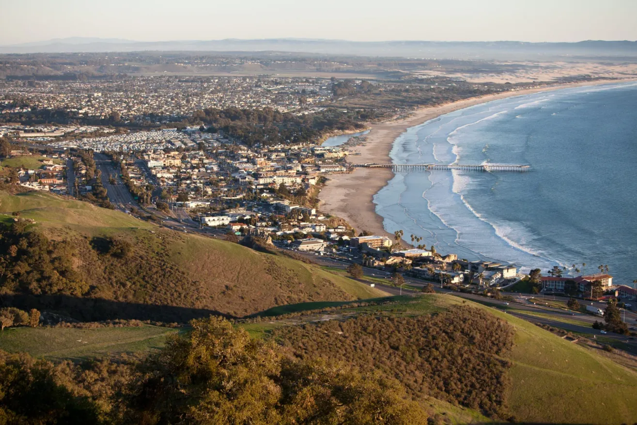 A coastal city with a long pier extends into the ocean, bordered by a wide sandy beach and a stretch of green hills in the foreground, under a clear sky.
