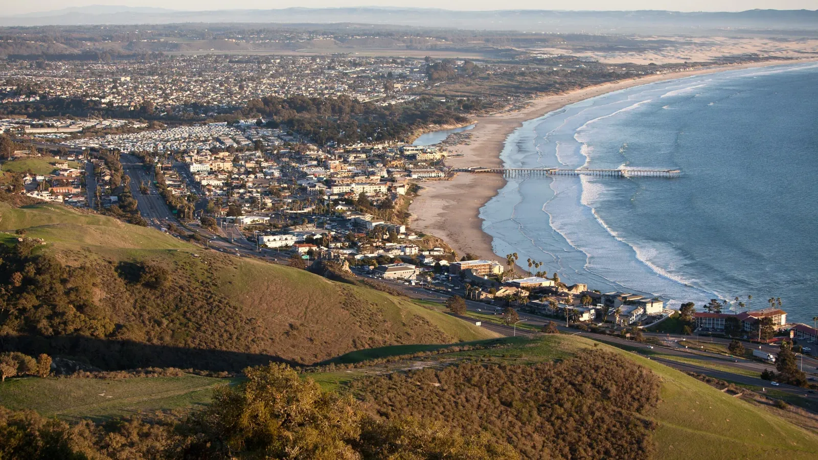 A coastal city with a long pier extends into the ocean, bordered by a wide sandy beach and a stretch of green hills in the foreground, under a clear sky.