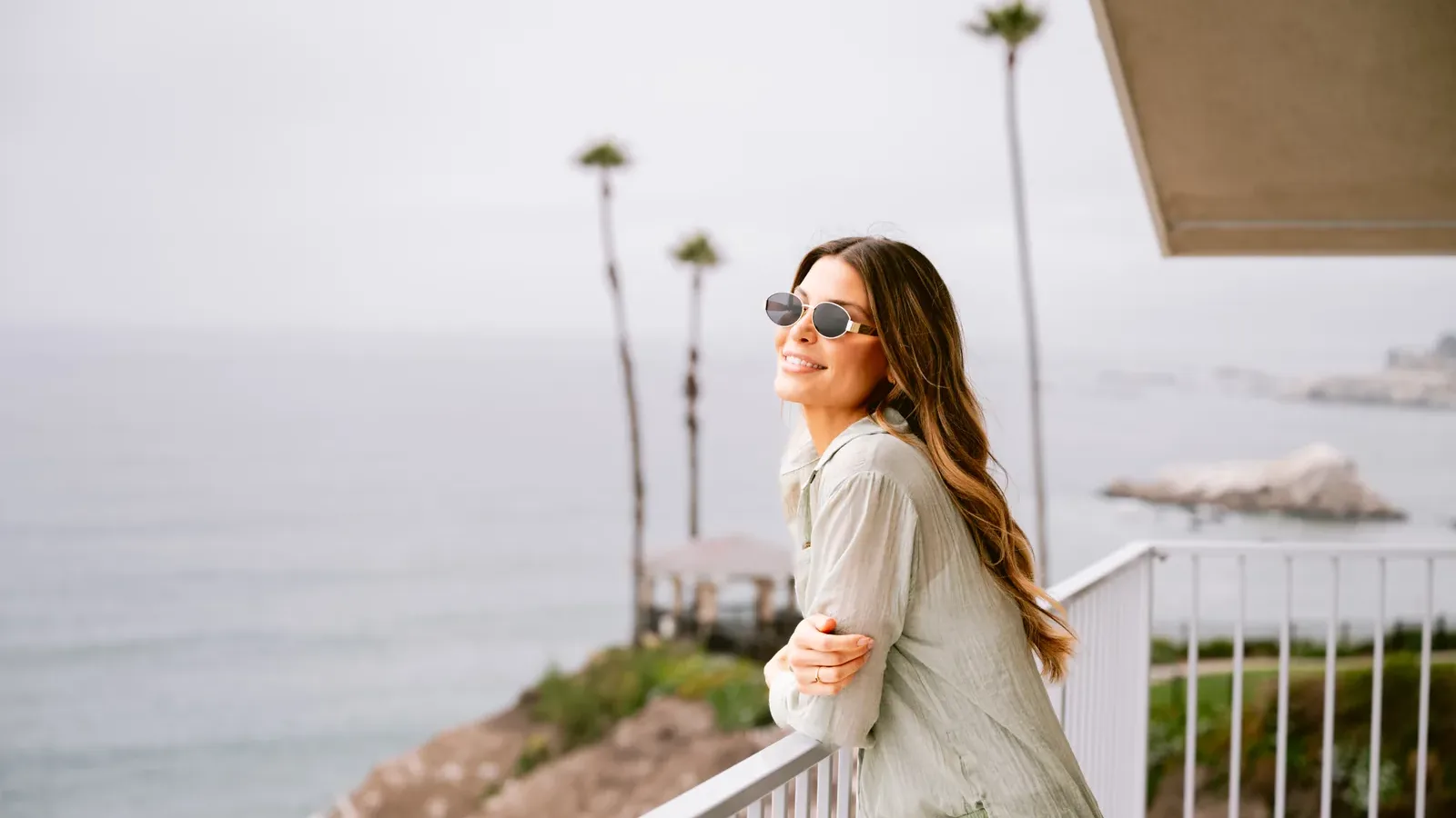 A woman in sunglasses and light clothing smiles while leaning on a balcony railing overlooking the ocean, with palm trees and a cloudy sky in the background.