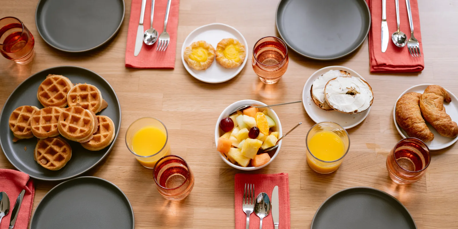 A breakfast table at Pismo Beach Resorts is set with gray plates, red napkins, utensils, glasses of orange juice, waffles, fruit salad, pastries, a bagel with cream cheese, and a croissant on a wooden surface.