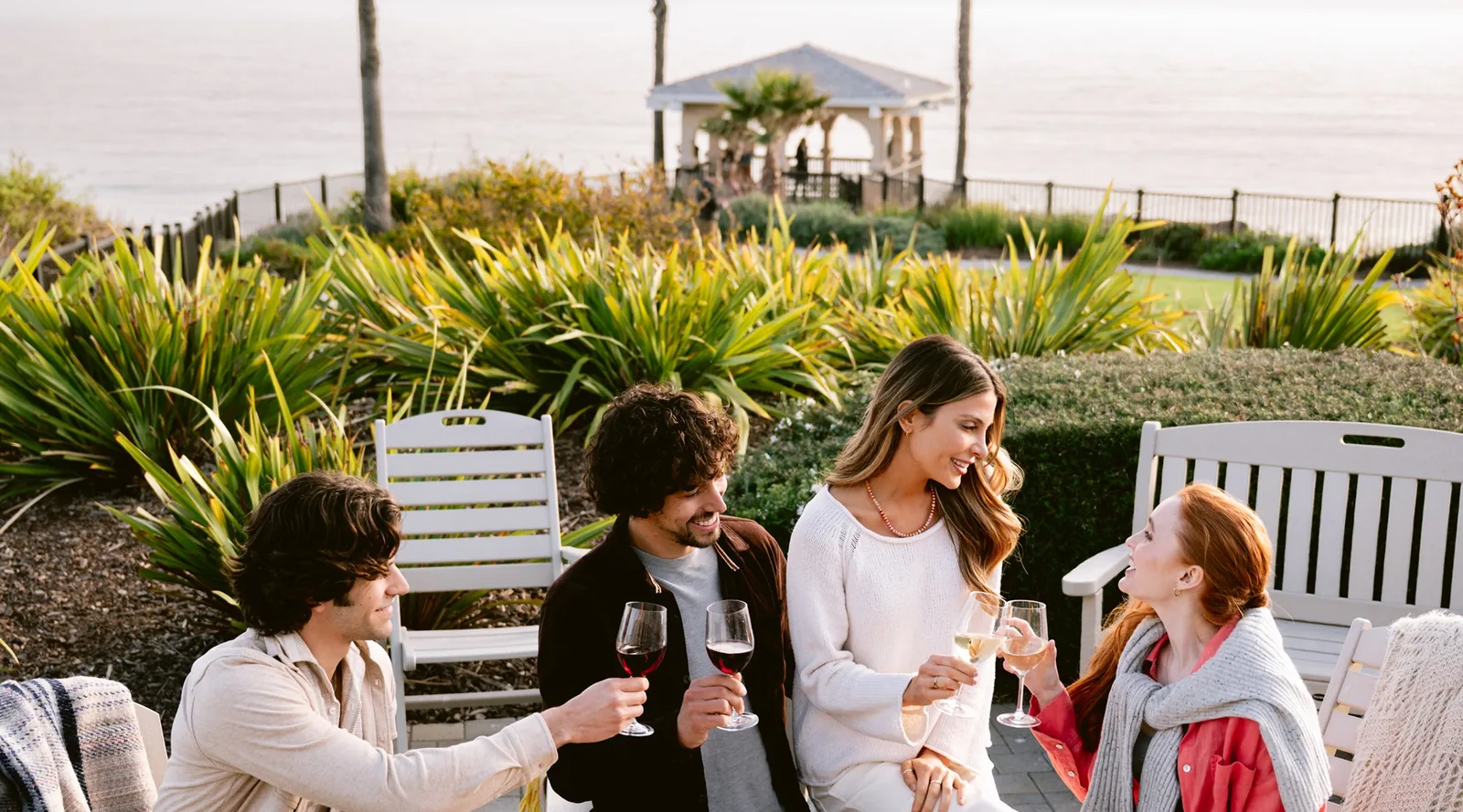 Four friends sit outdoors near greenery and toast with wine glasses, smiling and enjoying each others company. The ocean and a white gazebo are visible in the background.