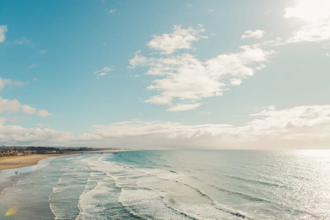 Wide sandy beach with gentle waves rolling onto the shore under a bright blue sky with scattered clouds. Small groups of people walk along the shoreline, and sunlight sparkles on the water.