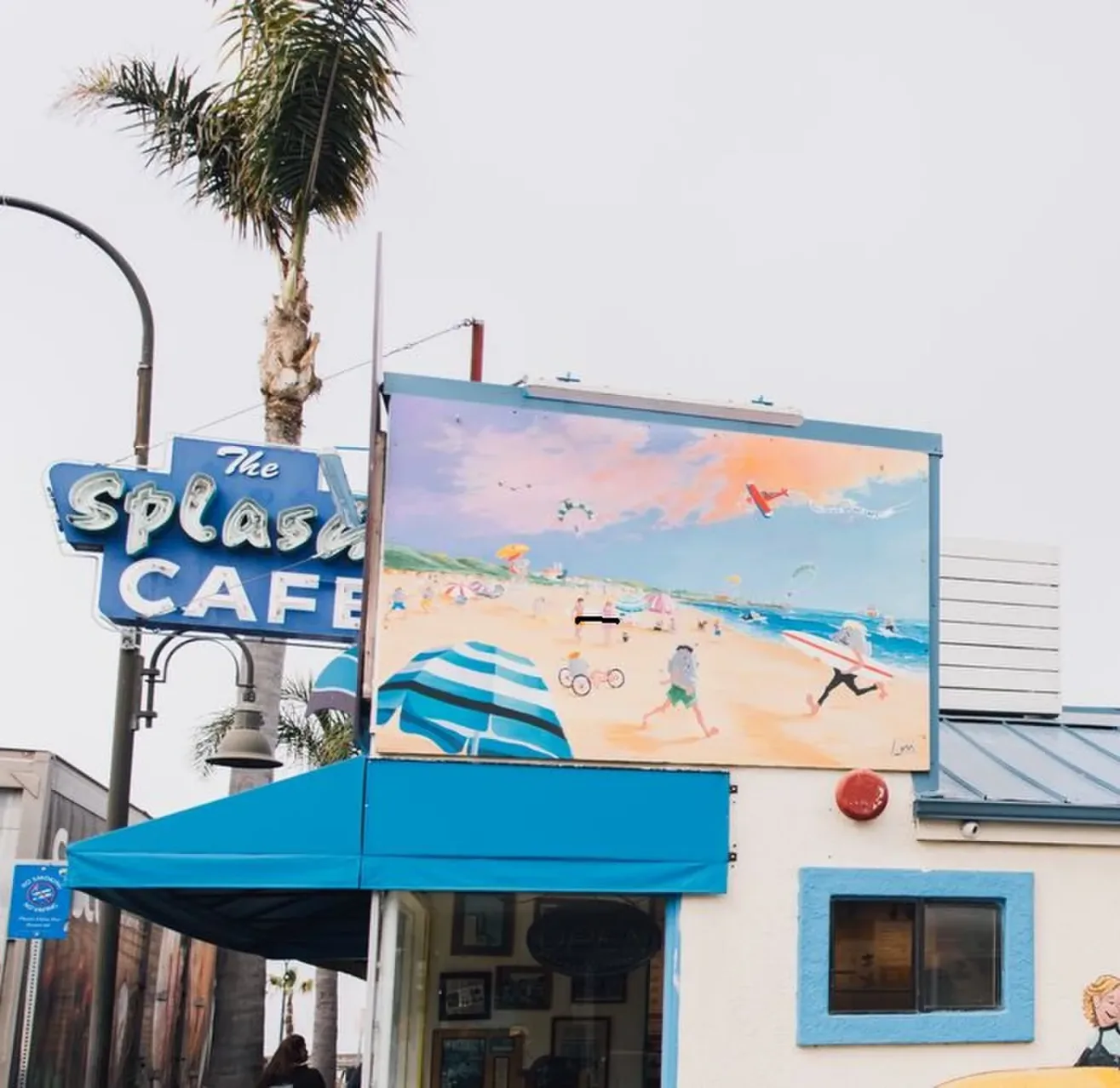 A colorful beach mural is displayed above the entrance of The Splash Café, which has a retro blue sign and a blue awning. A palm tree and streetlight are visible in the background under a cloudy sky.