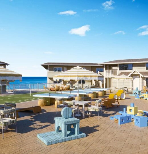 A spacious outdoor patio with tables, chairs, sun umbrellas, and childrens play equipment, overlooking a pool and a two-story building with the ocean visible in the background under a clear blue sky.