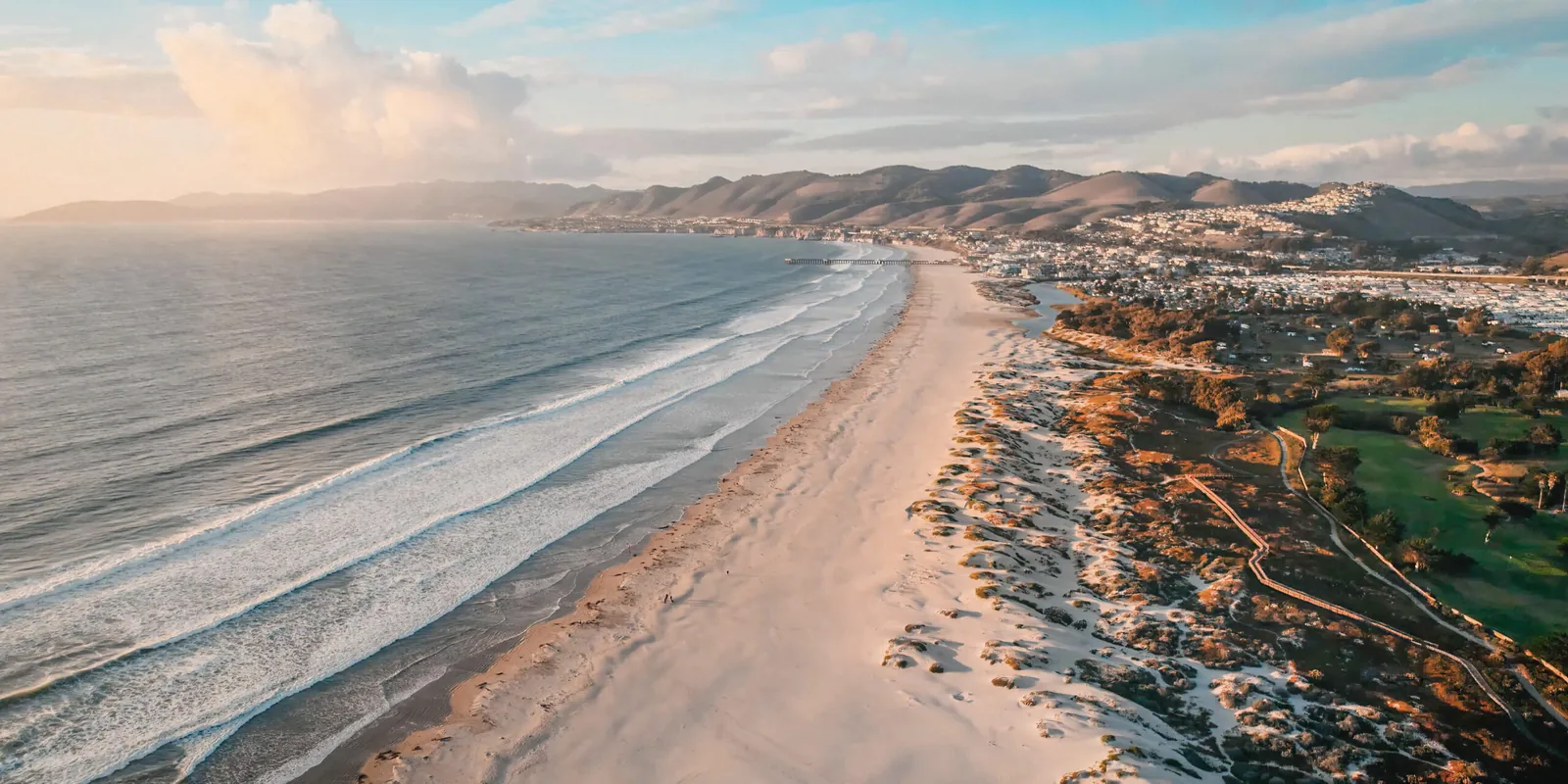 Aerial view of a wide sandy beach with gentle waves meeting the shore, a coastal town, rolling hills, and scattered trees under a partly cloudy sky at sunset.