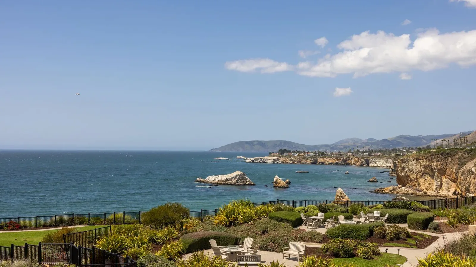 A coastal view with landscaped gardens, patio seating, and fences in the foreground overlooking the ocean, rocky shoreline, and distant hills under a blue sky with scattered clouds.