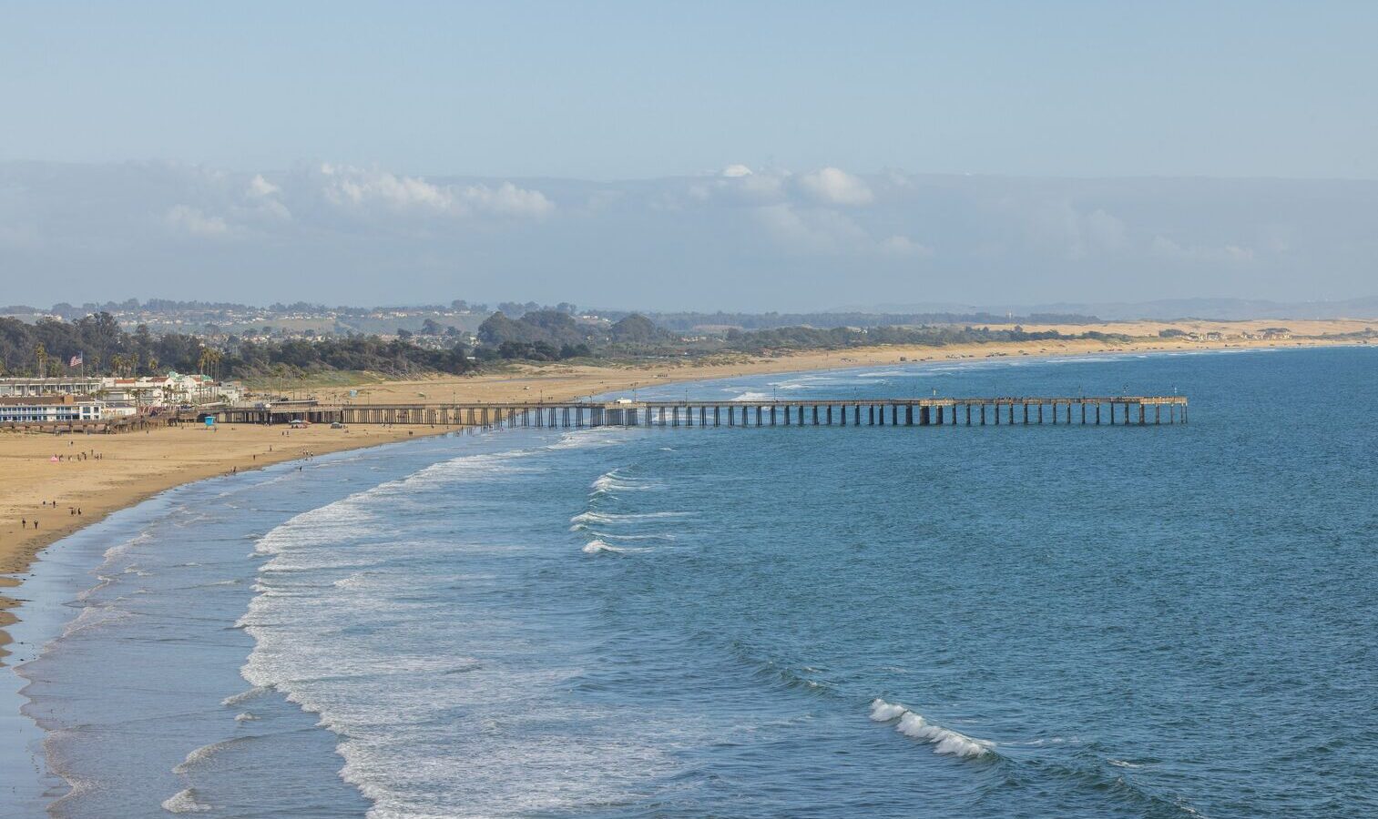 A long pier extends over the gentle waves of a wide sandy beach, with a coastal town and rolling hills visible in the background under a blue sky with scattered clouds.