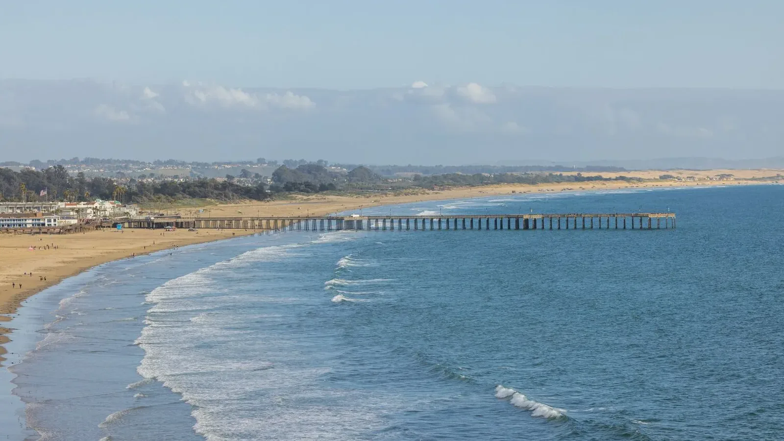 A long pier extends over the gentle waves of a wide sandy beach, with a coastal town and rolling hills visible in the background under a blue sky with scattered clouds.