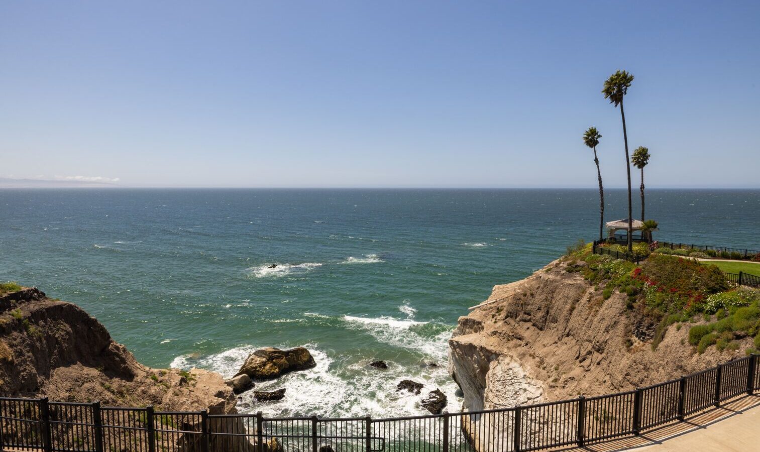 A coastal cliff with a black metal fence overlooks the ocean. Waves crash against rocks below, and a few tall palm trees stand on the cliff near a small shaded gazebo under a clear blue sky.