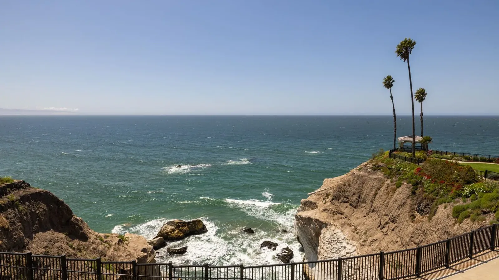 A coastal cliff with a black metal fence overlooks the ocean. Waves crash against rocks below, and a few tall palm trees stand on the cliff near a small shaded gazebo under a clear blue sky.