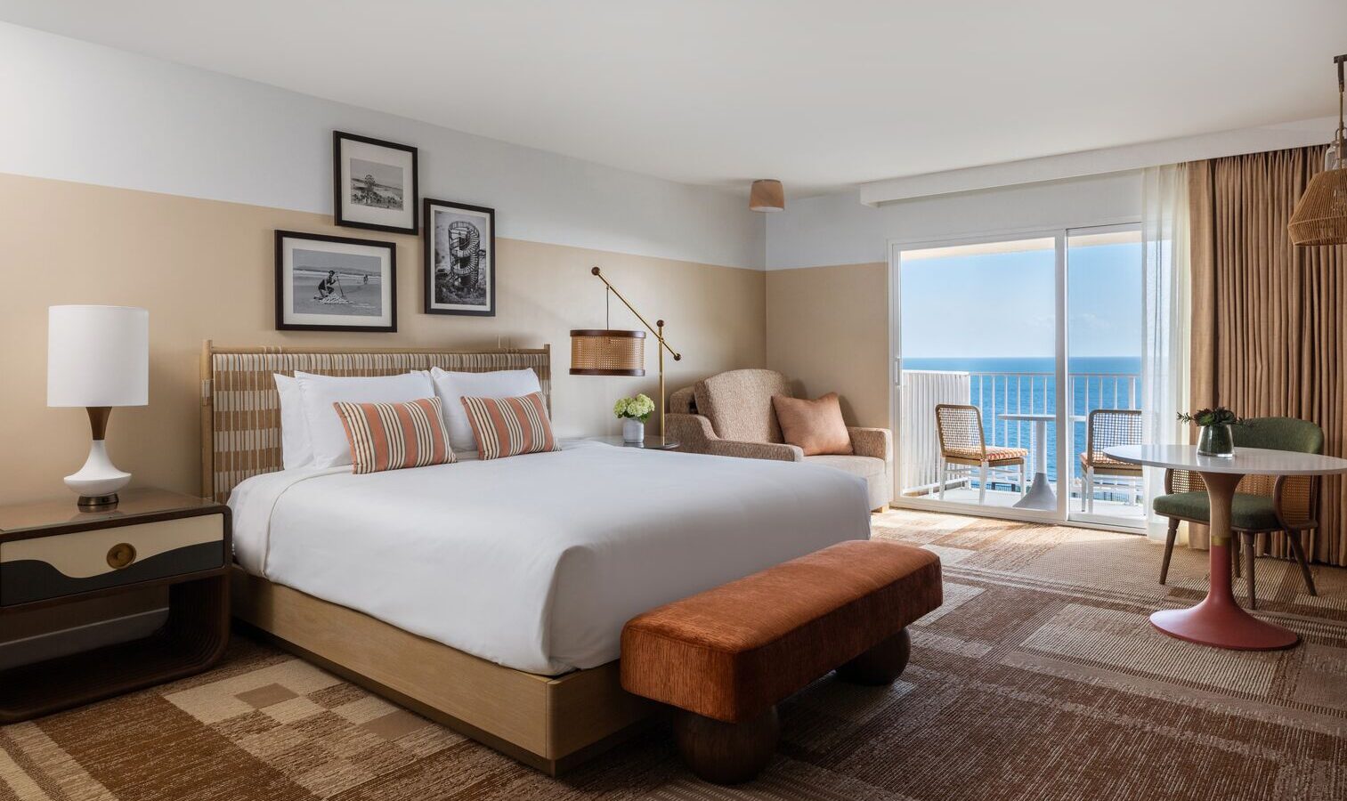 A modern hotel room with a large bed, striped pillows, nightstand, lamp, armchair, round table with chairs, artwork on the wall, and a balcony overlooking the ocean under a bright sky.