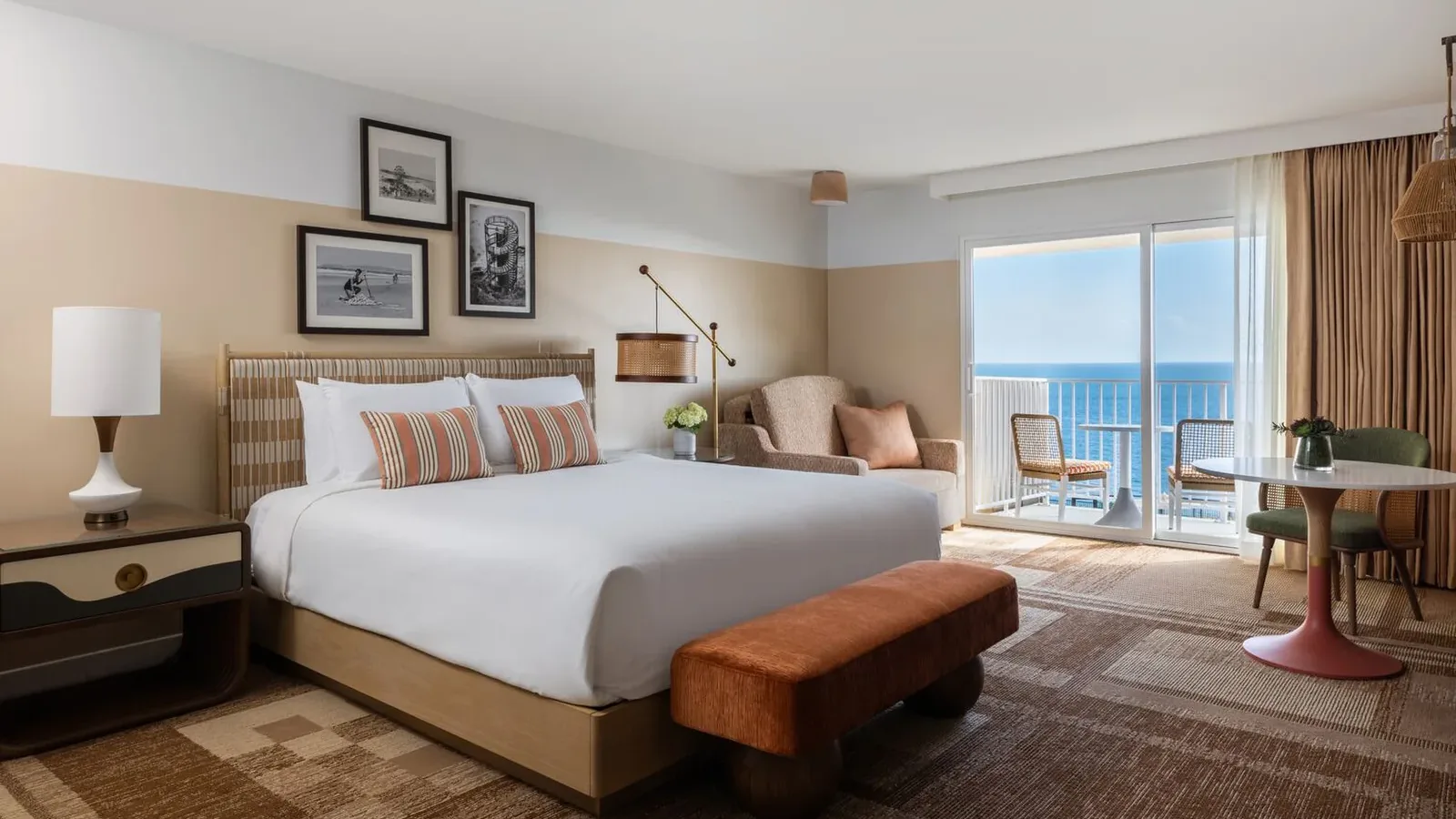 A modern hotel room with a large bed, striped pillows, nightstand, lamp, armchair, round table with chairs, artwork on the wall, and a balcony overlooking the ocean under a bright sky.