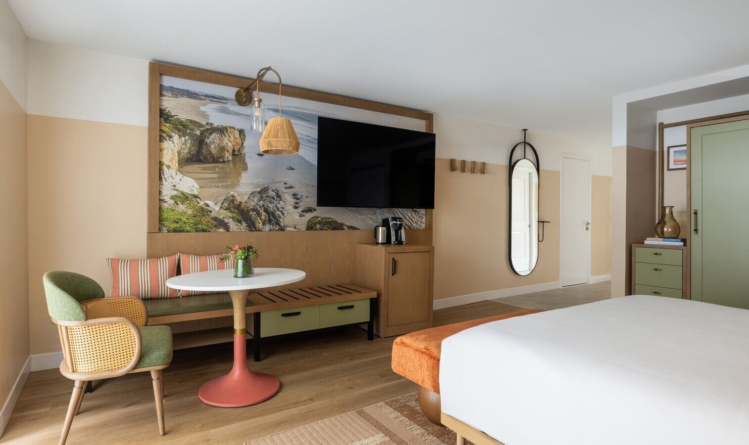 A modern hotel room with a bed, green and beige chairs, a round table, a wall-mounted TV, a large scenic photo, a long bench, a tall mirror, and minimalist decor in soft, neutral colors.