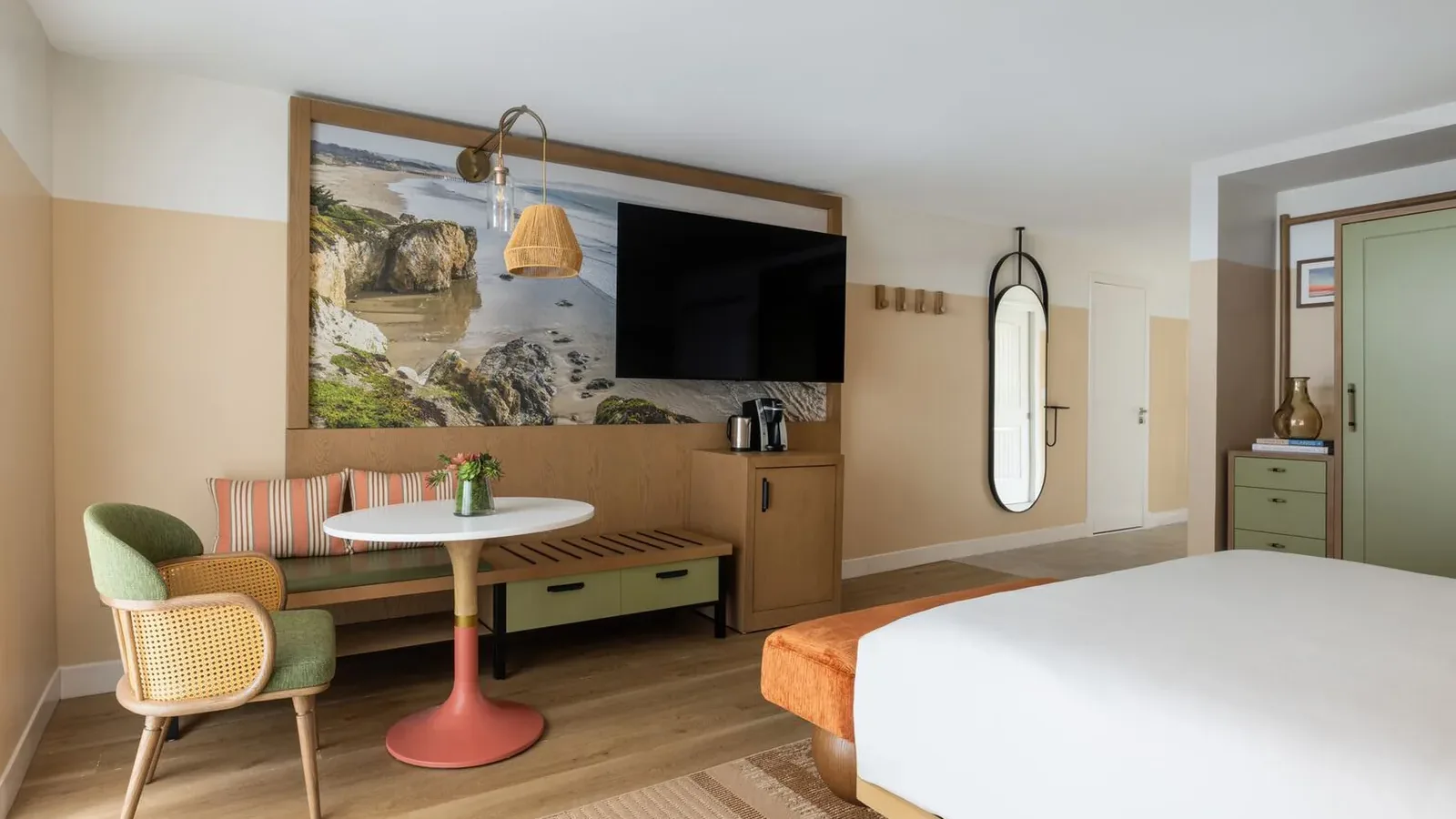A modern hotel room with a bed, green and beige chairs, a round table, a wall-mounted TV, a large scenic photo, a long bench, a tall mirror, and minimalist decor in soft, neutral colors.