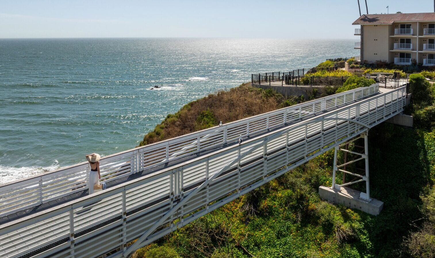 A metal walkway on stilts leads over green shrubs toward a seaside cliff, with an ocean view and a building with balconies on the right under a clear blue sky.