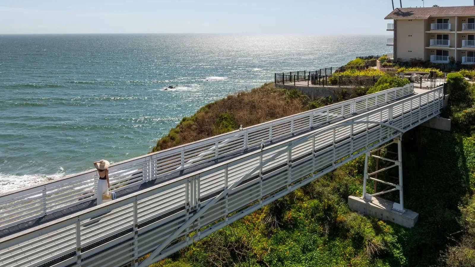 A metal walkway on stilts leads over green shrubs toward a seaside cliff, with an ocean view and a building with balconies on the right under a clear blue sky.