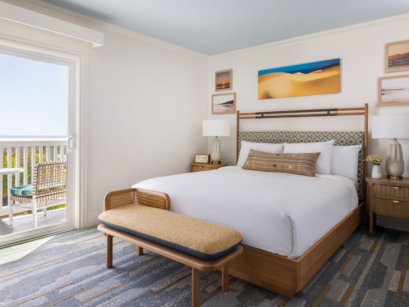 A bright, modern hotel room with a large bed, wooden bench, nightstands with lamps, framed art on the wall, and a sliding glass door leading to a balcony with outdoor seating and an ocean view.