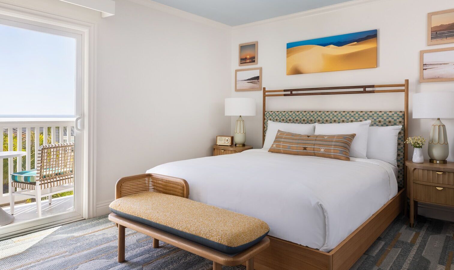 A bright, modern hotel room with a large bed, wooden bench, nightstands with lamps, framed art on the wall, and a sliding glass door leading to a balcony with outdoor seating and an ocean view.