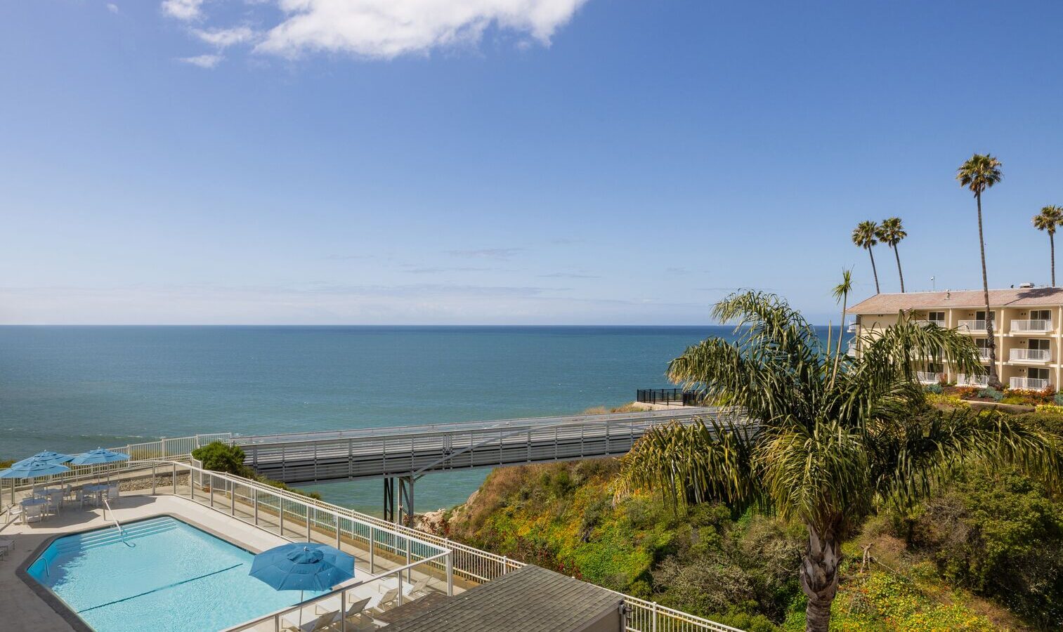 View of a turquoise swimming pool with blue umbrellas overlooking the ocean. A pier stretches over the water, and a hotel building with palm trees stands on a cliff to the right under a clear blue sky.