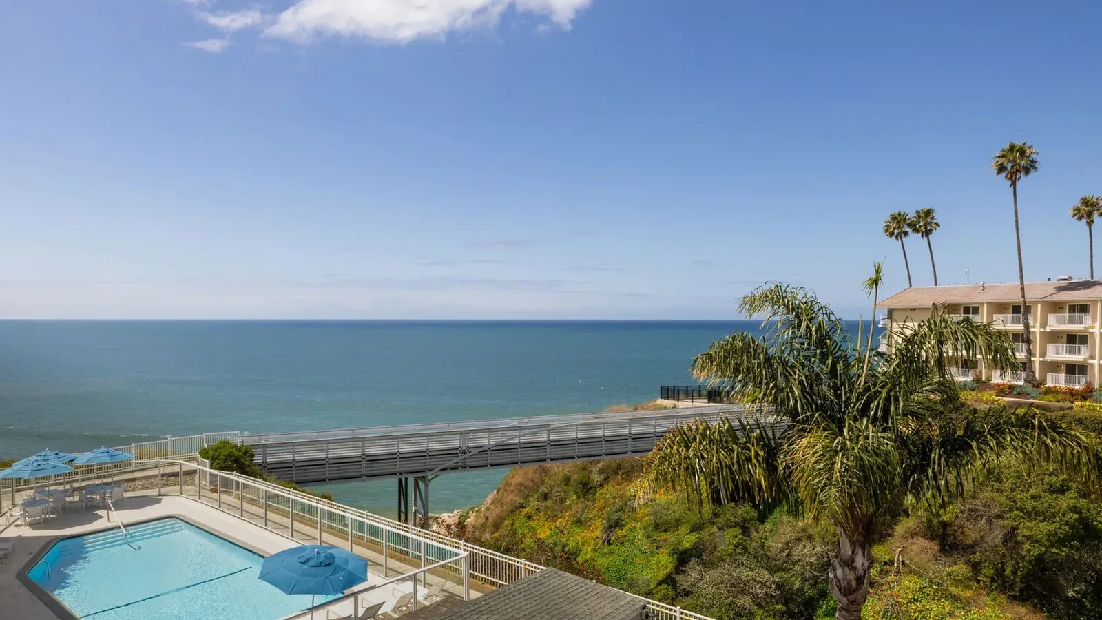 View of a turquoise swimming pool with blue umbrellas overlooking the ocean. A pier stretches over the water, and a hotel building with palm trees stands on a cliff to the right under a clear blue sky.