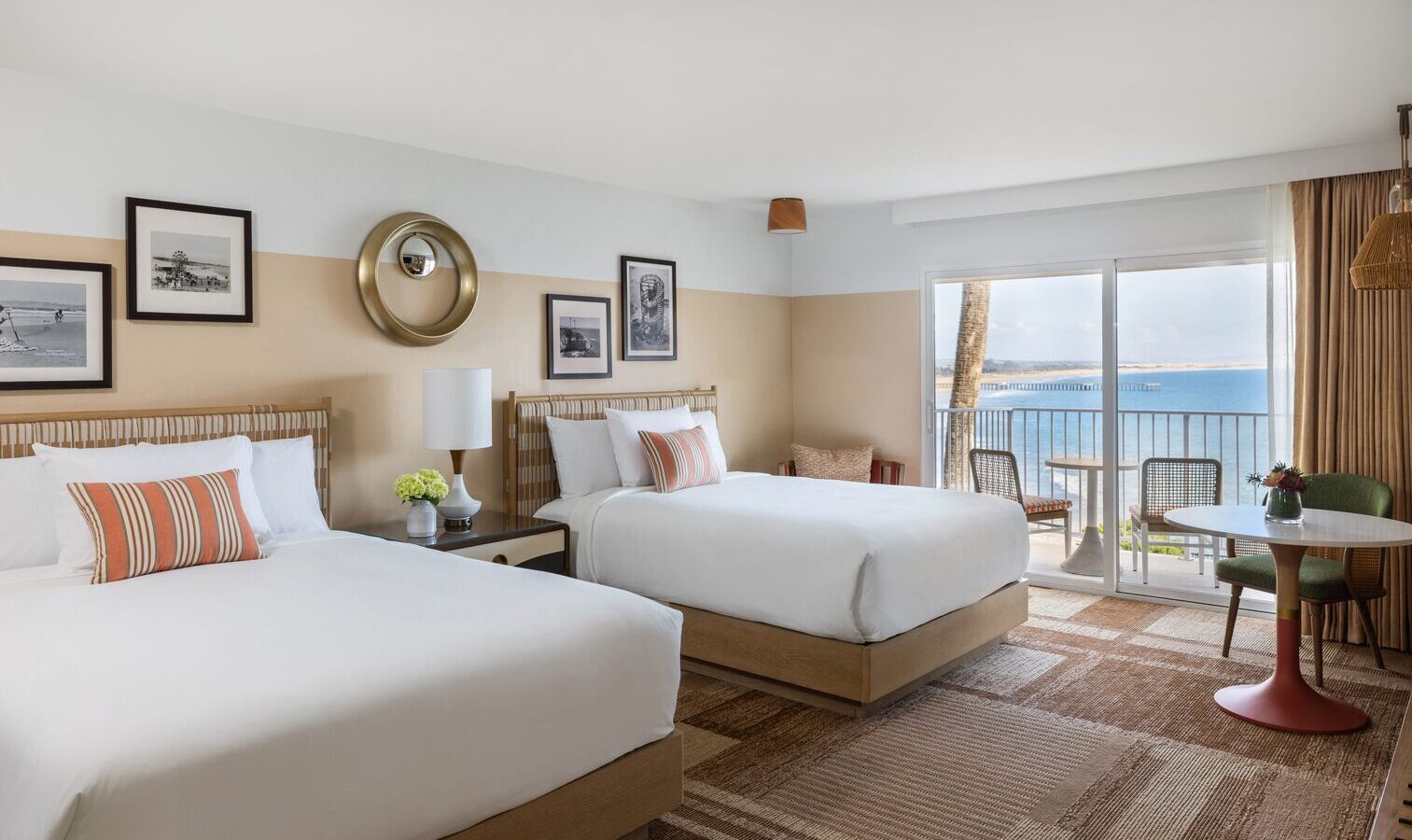 A bright hotel room with two double beds, striped pillows, framed art on the walls, a small round table with chairs, and a balcony overlooking the ocean under a clear sky.