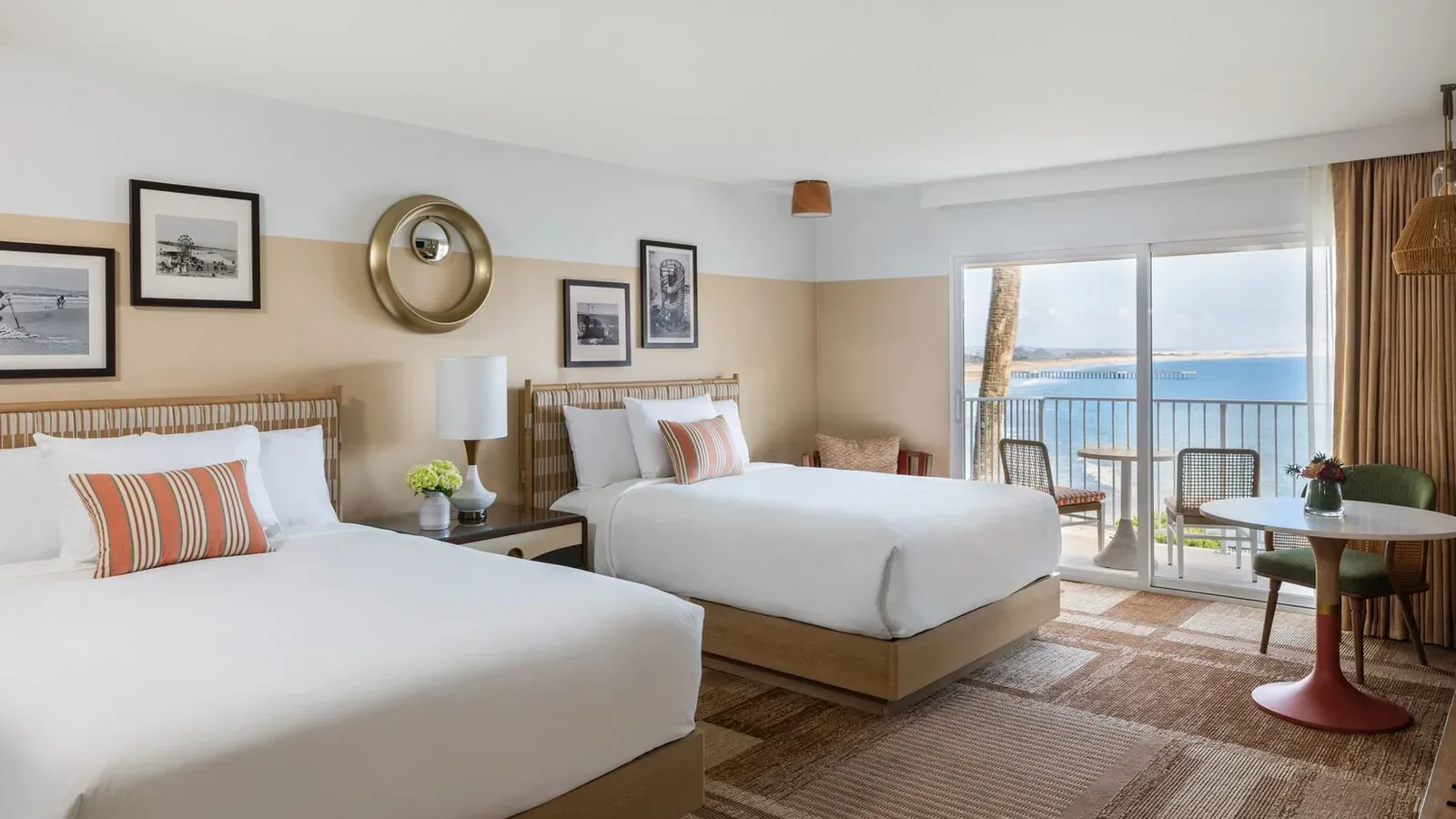 A bright hotel room with two double beds, striped pillows, framed art on the walls, a small round table with chairs, and a balcony overlooking the ocean under a clear sky.