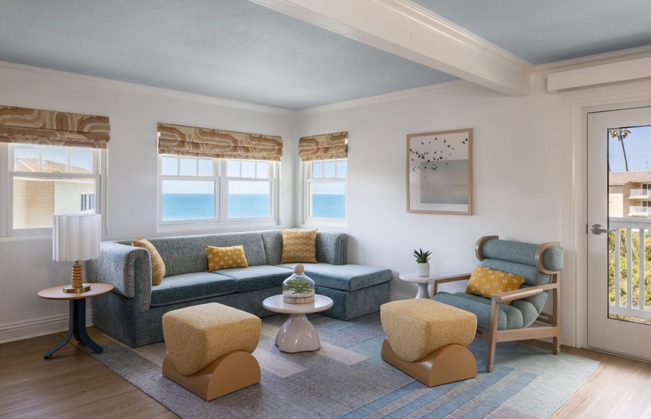 A bright, modern living room with large windows showing an ocean view, blue sectional sofa, yellow armchairs and ottomans, a round coffee table, a potted plant, wall art, and natural light.