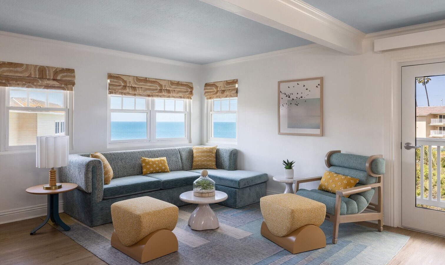 A bright, modern living room with large windows showing an ocean view, blue sectional sofa, yellow armchairs and ottomans, a round coffee table, a potted plant, wall art, and natural light.