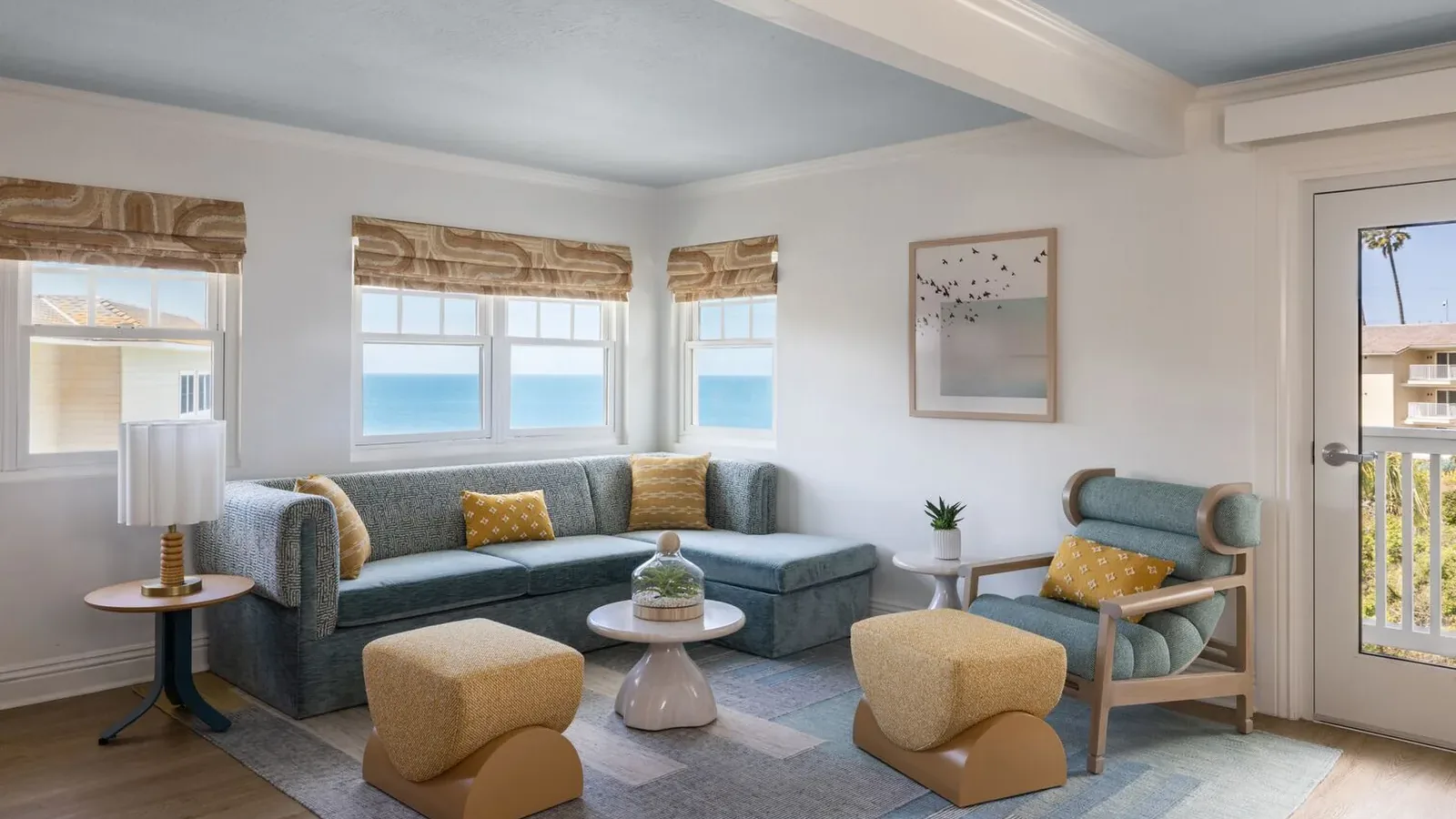 A bright, modern living room with large windows showing an ocean view, blue sectional sofa, yellow armchairs and ottomans, a round coffee table, a potted plant, wall art, and natural light.