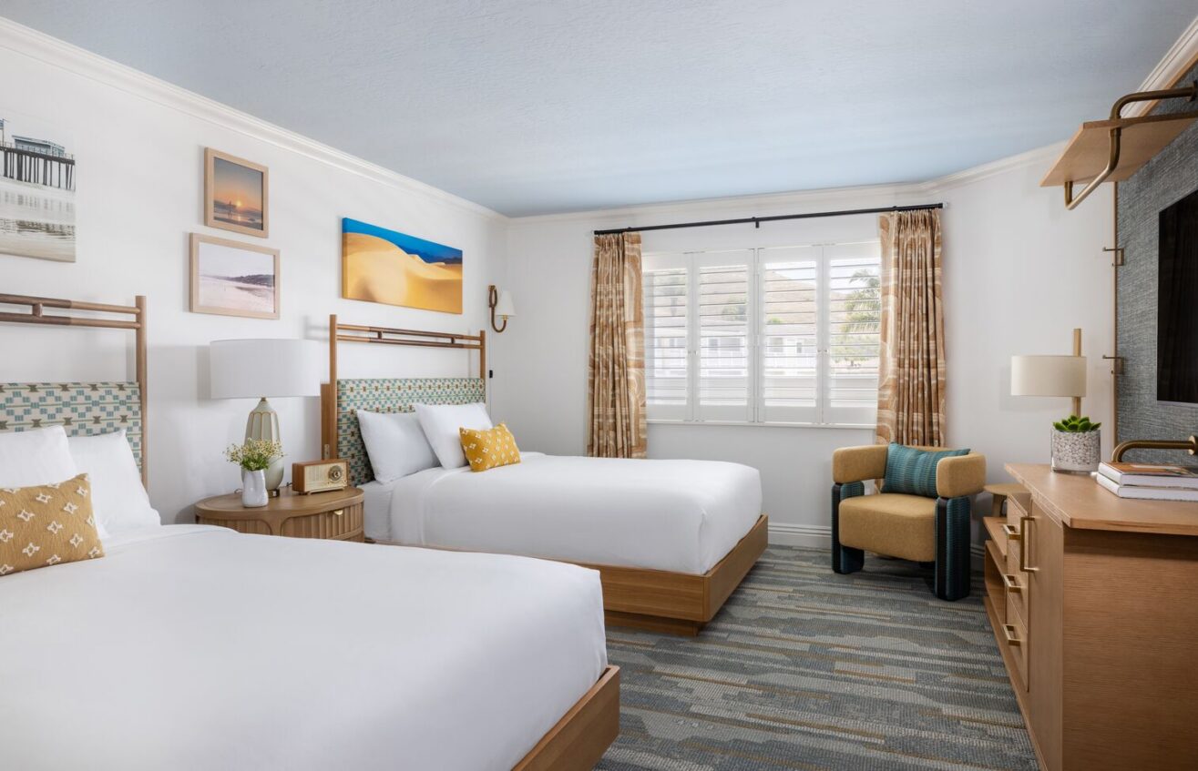 A bright hotel room with two double beds, wooden furniture, wall art, a TV, an armchair, and a window with shutters and beige curtains letting in natural light.