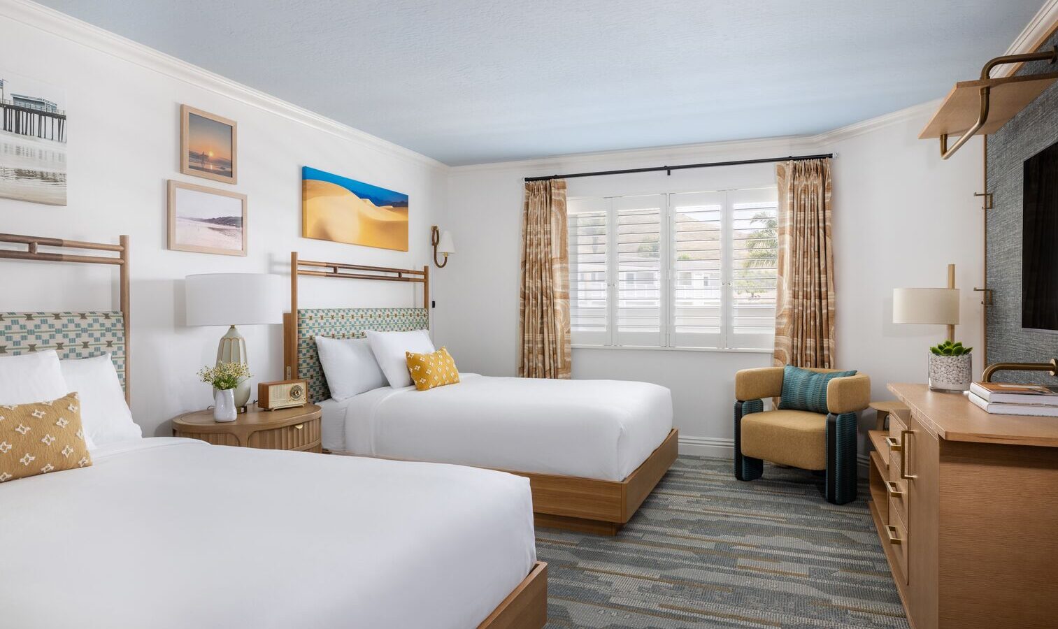 A bright hotel room with two double beds, wooden furniture, wall art, a TV, an armchair, and a window with shutters and beige curtains letting in natural light.
