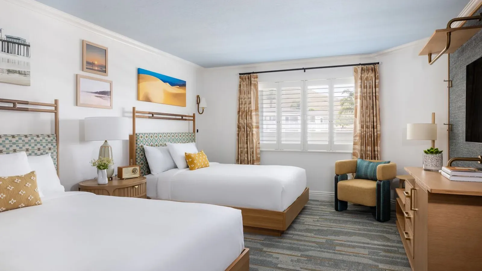 A bright hotel room with two double beds, wooden furniture, wall art, a TV, an armchair, and a window with shutters and beige curtains letting in natural light.